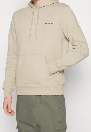 Beżowa bluza Carhartt z kapturem, z przednią kieszenią i sznurkami, noszona z oliwkowozielonymi spodniami typu cargo, z ręką wsuniętą w kieszeń.