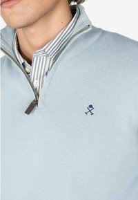 Jasnoniebieski sweter z dzianiny z zamkiem przy kołnierzu i brązowym uchwytem zamka. Na piersi haftowane granatowe logo. Nałożony na koszulę w paski.