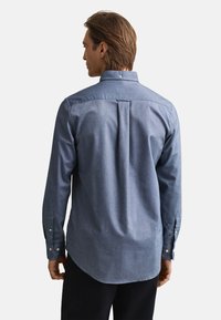 Zils chambray krekls ar pogājamu apkakli, garām piedurknēm un noapaļotu apakšu. Aprīkots ar aizmugurējo joslu un kasti ar krokām ērtākai kustībai un labākai piegādei.