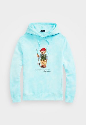 Lyseblå hættetrøje med et grafisk motiv af en fiskebjørn iført en rød kasket, grøn vest og beige bukser i midten, med teksten Polo Bear af Ralph Lauren nedenunder.