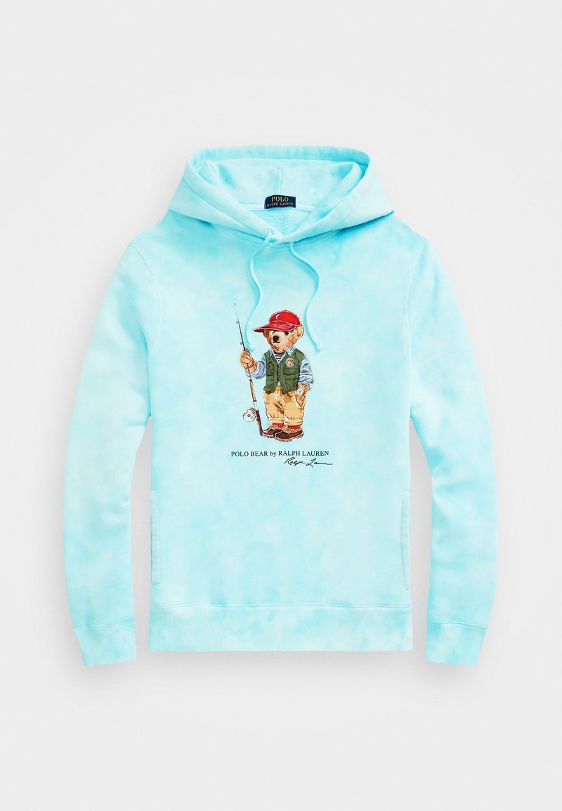 Sweat à capuche bleu clair avec un graphique d’un ours pêcheur portant une casquette rouge, un gilet vert et un pantalon beige au centre, avec le texte Polo Bear by Ralph Lauren en dessous.