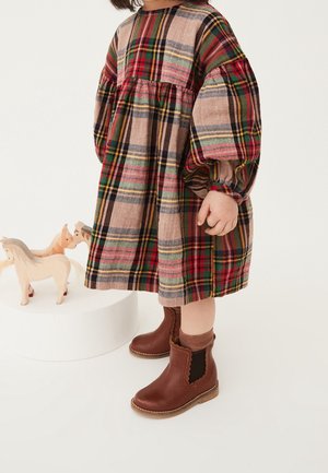 Robe à carreaux avec des manches bouffantes dans des tons beige, rouge et vert, associée à des bottes en cuir marron à la cheville et des chaussettes côtelées marron. Chevaux en bois à proximité.