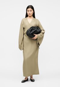 Femme debout portant une longue robe beige à larges manches, tenant une grande pochette noire, portant des chaussures plates noires.