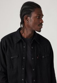 Camisa de ganga preta com pala de estilo western, botões de pressão, dois bolsos no peito e textura macia. Usada sobre uma t-shirt preta.