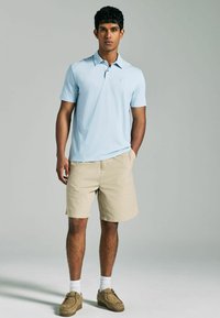 Hellblaues Poloshirt mit kurzen Ärmeln und Kragen, kombiniert mit beigen Shorts. Das Modell steht und trägt beige Schuhe und weiße Socken. Einfaches Design.