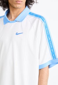 Polo branco da Nike com colarinho azul claro e mangas listradas. Apresenta um logotipo azul bordado e um padrão de riscas verticais subtil.