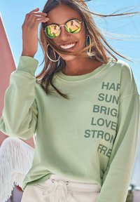 Hellgrüner Pullover mit dem Text "HARD SUN BRIGHT LOVE STRONG FREE" in dunklerem Grün, kombiniert mit silbernen runden Sonnenbrillen und weißen Hosen.