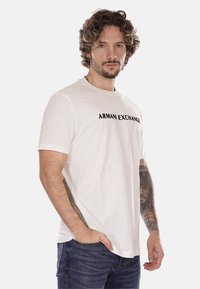 T-shirt bianco in cotone con la scritta "ARMANI EXCHANGE" stampata in lettere nere sul petto; presenta un classico collo rotondo e maniche corte.