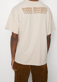 Béžové bavlněné tričko s oversized střihem, s opakujícím se textem "šťastné myšlenky" v tmavším odstínu na zadní straně. Krátké rukávy a kulatý výstřih.