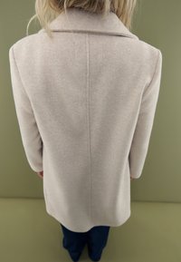 Abrigo de mezcla de lana color beige con cuello de muesca, corte recto, mangas largas y detalles de costura sutiles, sobre un fondo verde.