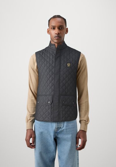 Veste matelassée noire avec un col montant, deux poches avant et un logo doré. Portée sur une chemise à manches longues beige avec un jean bleu.