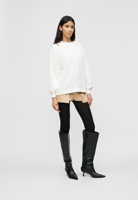 Γυναίκα που στέκεται φορώντας λευκό φαρδύ πουλόβερ, μπεζ σορτς, μαύρο κολάν και μαύρες μπότες με τακούνι μέχρι το γόνατο, μπροστά σε απλό φόντο.