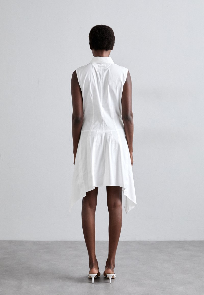 Hvid ærmeløs kjole med asymmetrisk nederkant, lavet af letvægtsstof, med krave og knapdetalje på ryggen.