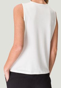 Haut sans manches blanc avec un col rond, texture lisse et coupe décontractée, associé à un pantalon sombre, vu de dos.