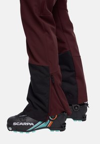 Heren ski-broek in bordeauxrood en zwart, met ritsen bij de manchetten en verstevigde onderzomen, ontworpen voor duurzaamheid en bewegingsvrijheid.