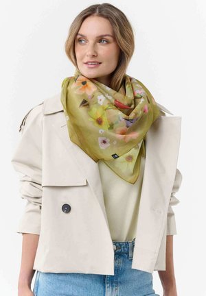 Woman wearing a light beige cropped trench coat, blue jeans, and a green floral scarf, looking slightly to the side with a neutral background.