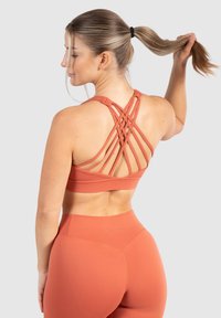 Sujetador deportivo con un diseño de espalda en forma de raqueta y tirantes cruzados, hecho de una tela suave y elástica en color terracota, combinado con leggings de cintura alta.