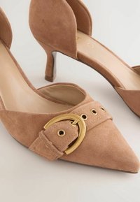 Escarpins en daim beige à bout pointu, ornés d'une boucle dorée décorative et dotés d'un petit talon kitten, sur un fond clair uni.