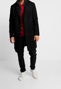 Mężczyzna w czarnym długim płaszczu, koszulce w czerwono-czarne tie-dye, czarnych spodniach i białych sneakersach stoi na białym tle.