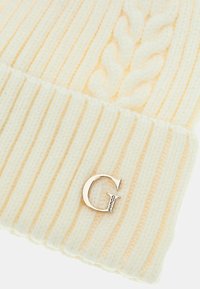 Bonnet tricoté jaune clair avec une texture côtelée et un motif en cable. Présente un accent en métal argenté avec le logo "G". Tissu doux et chaud.