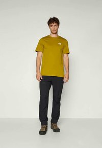 Camiseta de algodón verde oliva con mangas cortas y un logo en el pecho, combinada con pantalones negros y botas de senderismo marrones.