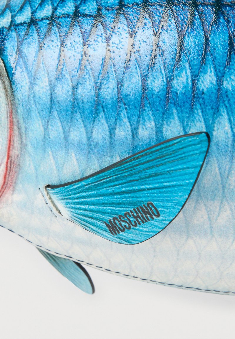 Gros plan sur le corps texturé bleu d'un poisson avec une nageoire portant l'inscription "MOSCHINO" sur un fond blanc.