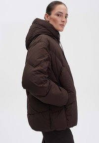 Giacca puffer marrone con cappuccio, caratterizzata da una texture trapuntata e un fit rilassato. La chiusura con zip e i lacci regolabili aggiungono funzionalità.