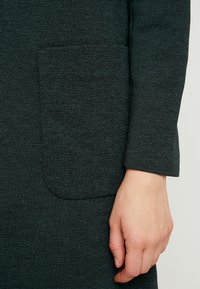 Mørkegrøn, tekstureret kjole med synlig lomme og lange ærmer. Stoffet har et ribbet mønster og en glat finish.