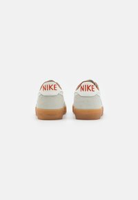 Adida�și Nike cu partea superioară din suede gri, tab de călcâi portocaliu și talpă din cauciuc natural. Prezintă logo-ul "NIKE" brodat în roșu pe călcâi.