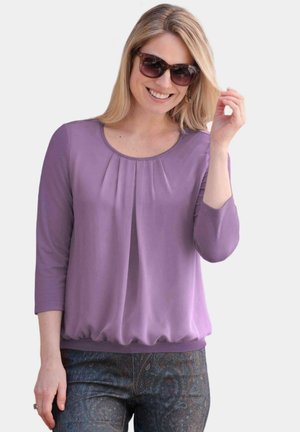 Lavendelfarbene Bluse mit Dreiviertelärmeln, gefältelter Vorderseite und elastischem Saum, kombiniert mit gemusterten dunklen Hosen. Model trägt Sonnenbrille.