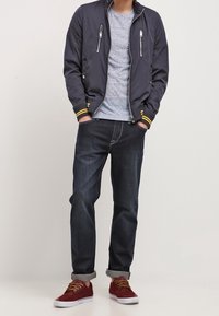 Giubbotto bomber navy con dettagli gialli, t-shirt grigia, jeans scuri arrotolati ai polsini e sneakers bordeaux. Tasche con zip sul giubbotto.