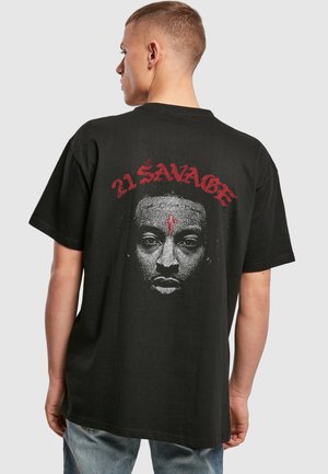 Jeune homme portant un t-shirt noir avec une grande illustration d'un visage et un texte rouge "21 Savage" dans le dos, debout devant un fond uni.