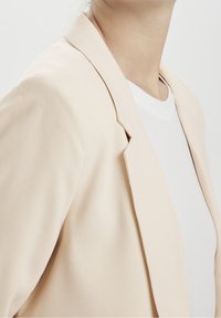 Beige blazer se strukturovaným designem a hladkou texturou, s klopou a draped přední částí, vrstvený přes bílou košili.