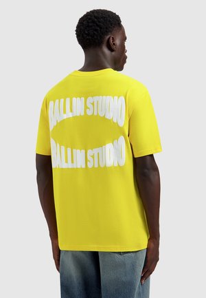 Man zijwaarts gezicht draagt een knalgele T-shirt met grote witte tekst "BALLIN STUDIO" twee keer op de achterkant, gecombineerd met een blauwe spijkerbroek.