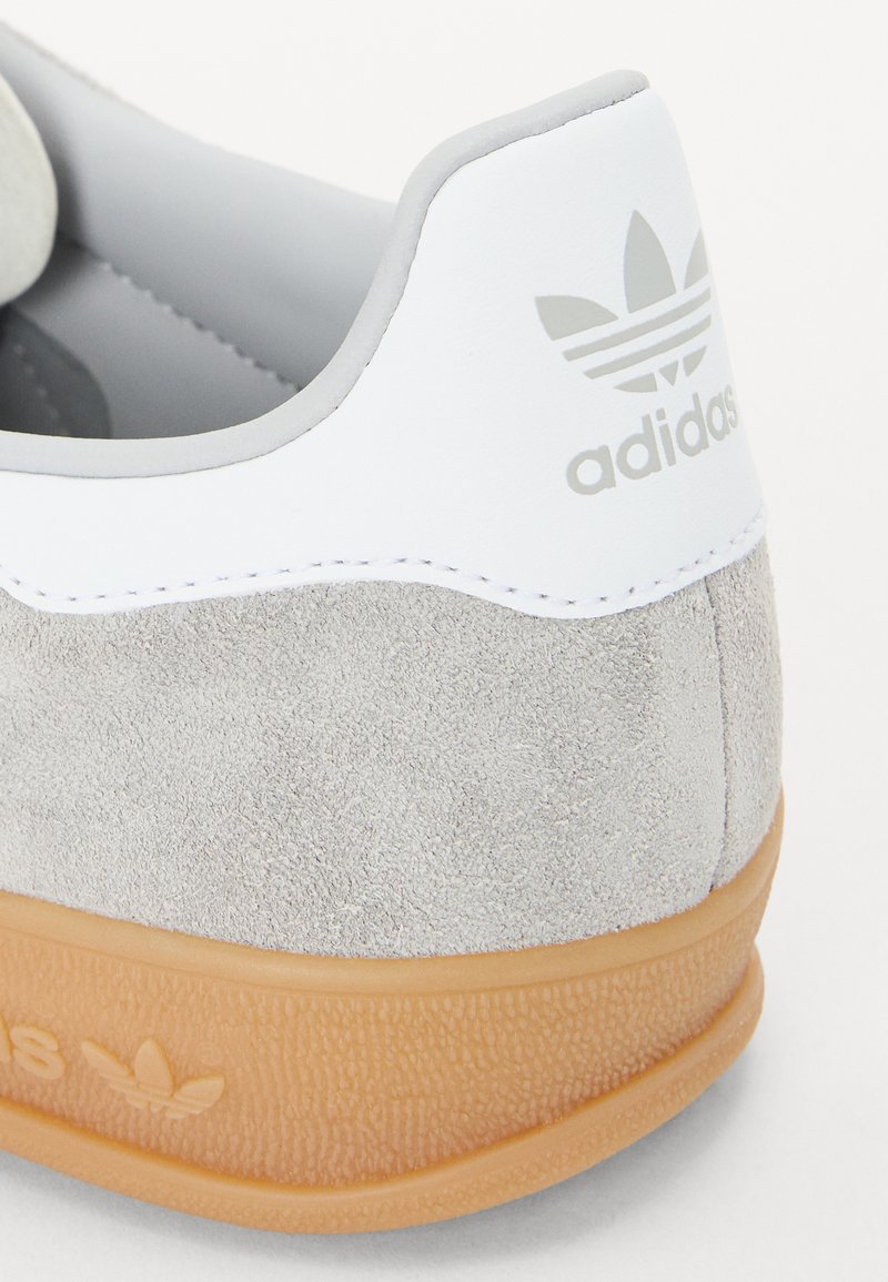Zapatillas de gamuza gris y cuero blanco con suela de goma, con el logo de Adidas grabado en el talón. Texturas suaves y líneas limpias.