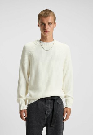 Hvid ribbet striktrøje med rund hals og sænkede skuldre, parret med mørkegrå løse jeans med et mærket sort bælte.