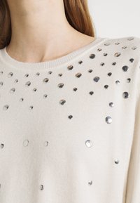 Hellbeige Pullover mit Rundhalsausschnitt und verstreuten silbernen Verzierungen, die eine strukturierte und visuell dynamische Oberfläche schaffen.
