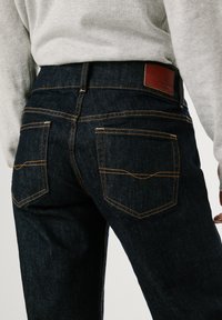 Temno modri traper hlače z oranžnim šivanjem, s svetlo rjavim usnjenim oznako na pasu in dvema zadnjima žepoma z naklonjenimi pokrovi.