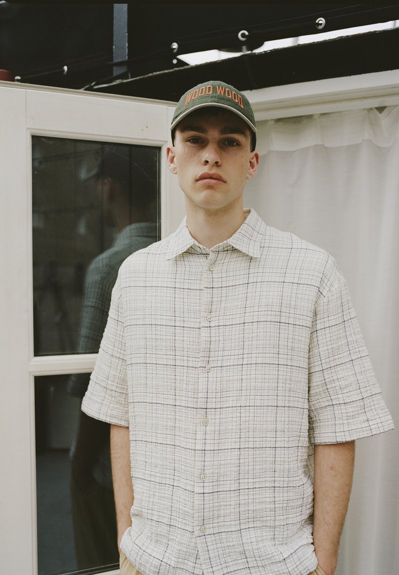 Chemise à manches courtes, boutonnée en crème avec un motif à carreaux. Portée avec une casquette de baseball verte arborant un texte orange. Tissu simple et texturé.