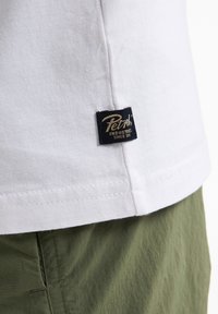 Witte katoenen t-shirt met een zwart label met gouden tekst "Petzl Industries Sinds 1973." Het t-shirt heeft een eenvoudige ontwerp en een afgeronde zoom.