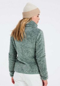 Giacca in pile verde fuzzy con colletto alto e maniche lunghe. Abbinata a un berretto lavorato a maglia color crema e beige. Tessuto morbido, vestibilità casual.
