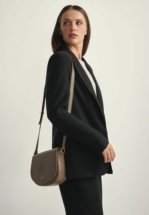 Kobieta w czarnej marynarce i spodniach, niosąca taupe skórzaną torbę na ramię, spoglądająca na bok na jednolitym jasnym tle.
