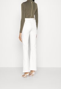 Top de manga larga con costillas en color oliva y cierre de cremallera en la espalda, combinado con pantalones acampanados blancos y sandalias blancas de tacón alto.