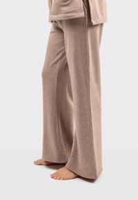 Jasnobrązowe spodnie typu wide-leg wykonane z miękkiego, teksturowanego materiału, z bocznymi szwami i luźnym krojem, noszone na boso na jednolitym tle.