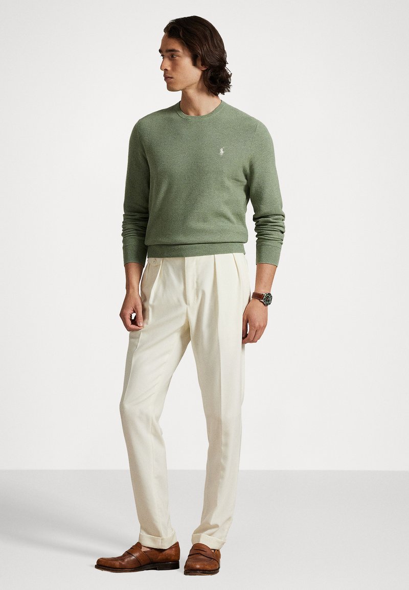Grüner Strickpullover mit rundem Ausschnitt, kombiniert mit cremefarbenen, gefalteten Hosen und braunen Slippern. Dezentes Logodetail auf dem Pullover.