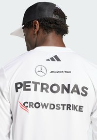 Jersey deportivo blanco con logotipos impresos en negro: Mercedes, AMG, Petronas, Crowdstrike; cuenta con un cuello redondo y mangas largas. Gorra negra con parte trasera de malla.