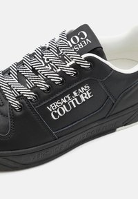 Sapatilha preta em pele com logotipo branco em contraste, atacadores às riscas e uma sola de borracha texturizada com detalhes de marcação na lateral.