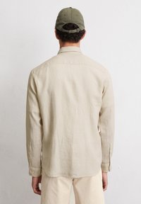 Beige langemouwen shirt met een rechte zoom en een zachte textuur, gecombineerd met crèmekleurige broek. Het model draagt een groene pet.