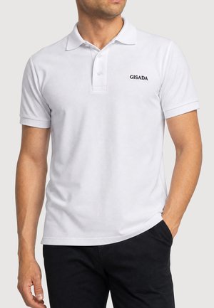 Mann trägt ein weißes kurzärmeliges Poloshirt mit "GISADA"-Logo und schwarze Hose, eine Hand in der Hosentasche, leicht nach links gewandt.