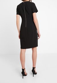 Schwarzes Kleid mit kurzen Ärmeln, figurbetontem Schnitt, mittellanger Höhe bis zur Mitte des Oberschenkels, mit einem auffälligen Reißverschluss auf der Rückseite und glatter Textur.